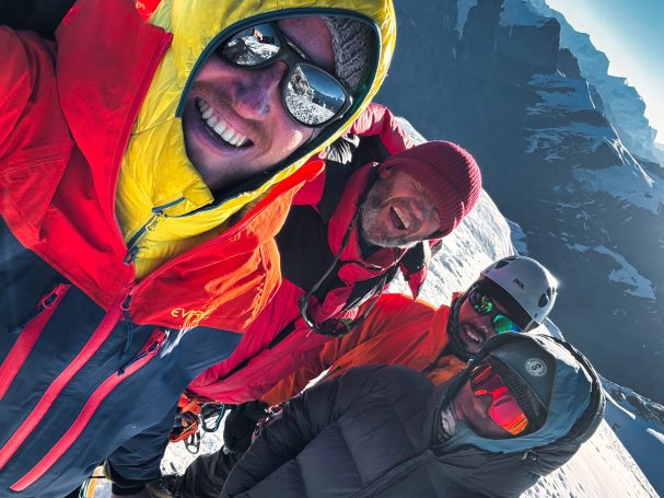 Vier Bergsteiger in farbiger Ausrüstung lächeln auf einem Gipfel mit schneebedeckter Landschaft.