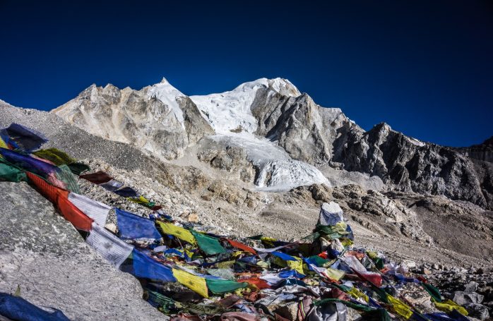 Bunte Gebetsfahnen liegen auf einem Gebirgspfad mit schneebedecktem Gipfel im Hintergrund.