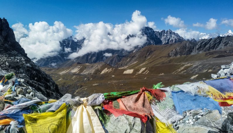 Berglandschaft mit bunten Gebetsfahnen, Wolken und schneebedeckten Gipfeln im Hintergrund.
