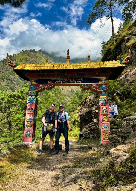 Zwei Wanderer stehen vor einem bunten Tor in einer bergigen, grünen Landschaft.