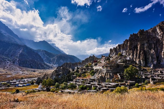 Landschaft mit Bergen, Kloster und grünen Feldern unter einem blauen Himmel.