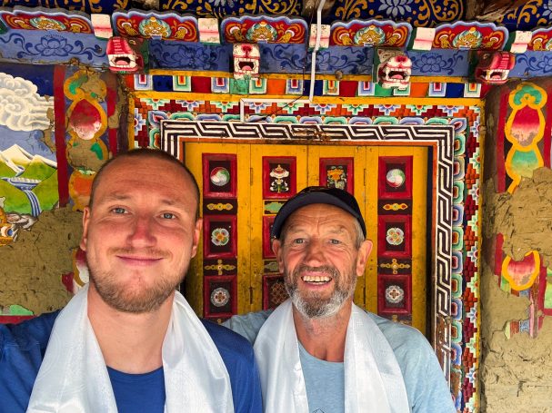 Zwei Männer lächeln vor einer farbenfrohen, kunstvoll dekorierten Tür.