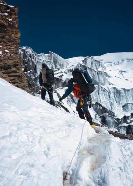 Zwei Bergsteiger auf einem schneebedeckten Hang mit Gletscher im Hintergrund.