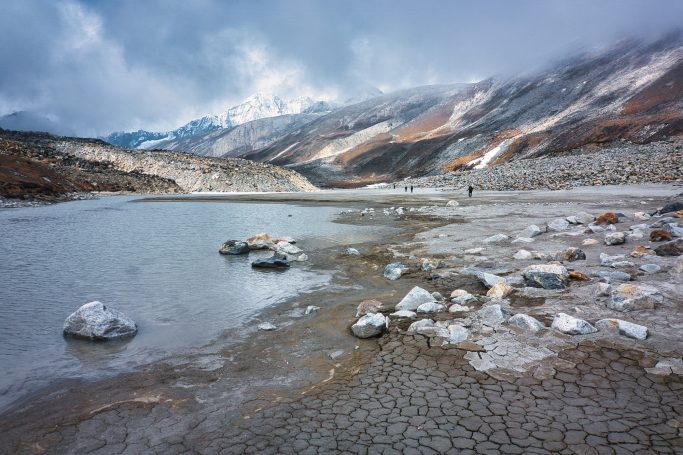 Berglandschaft mit See, Steinen und nebligem Himmel, im Hintergrund schneebedeckte Berge.
