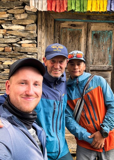 Drei Männer posieren lächelnd vor einer Holztür in einem Bergdorf.