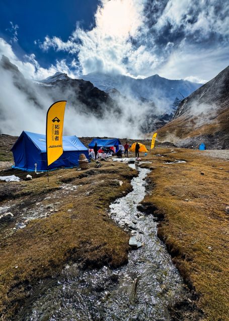 Zeltlager in den Bergen mit bunten Fahnen, umgeben von Nebel und einem kleinen Bach.