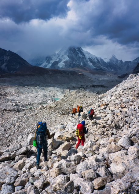Bergsteiger auf steinigem Gelände, umgeben von schneebedeckten Gipfeln und dunklem Himmel.