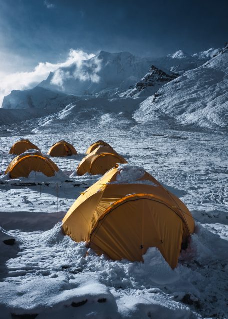 Gelbe Zelte im Schnee, umgeben von Bergen und bewölktem Himmel.