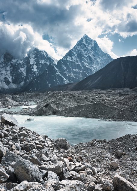 Berglandschaft mit schroffen Gipfeln, einem See und grauen Felsen unter bewölktem Himmel.