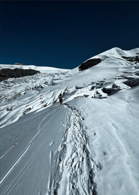 Schneebedeckter Bergweg mit spärlicher Vegetation und klarem blauen Himmel.