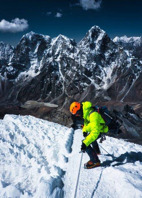 Bergsteiger in leuchtender Jacke auf schneebedecktem Gipfel mit hohen Bergen im Hintergrund.