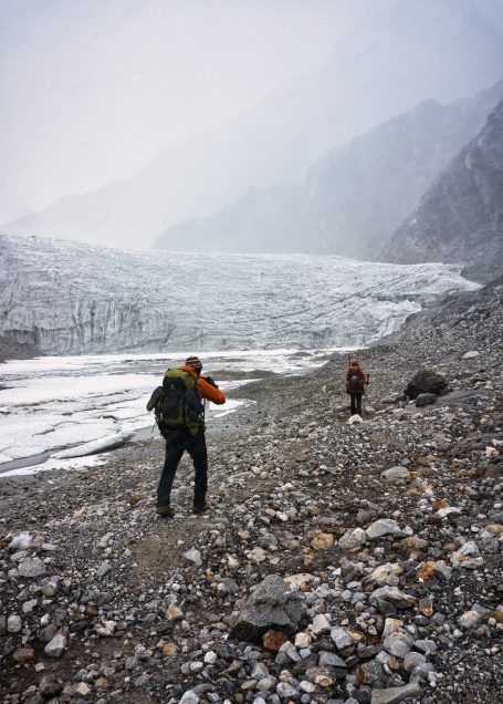Zwei Wanderer erkunden eine felsige, eisige Landschaft mit einem Gletscher im Hintergrund.