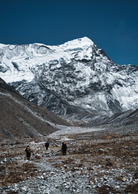 Drei Wanderer auf einem steinigen Weg mit schneebedecktem Berg im Hintergrund.
