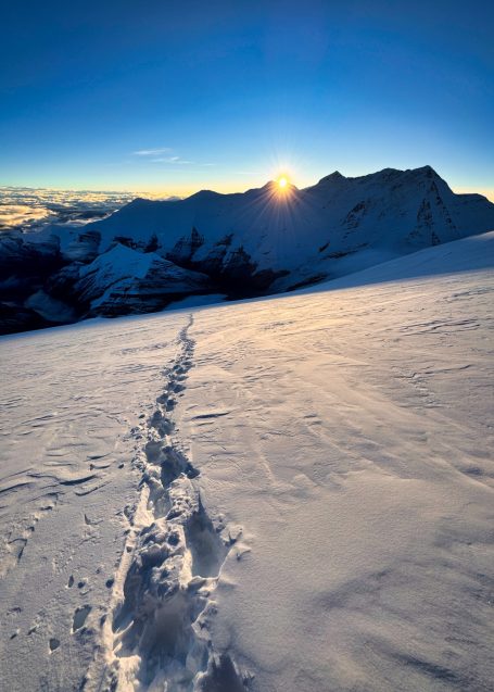 Spuren im Schnee unter einem Sonnenaufgang über Berggipfeln.