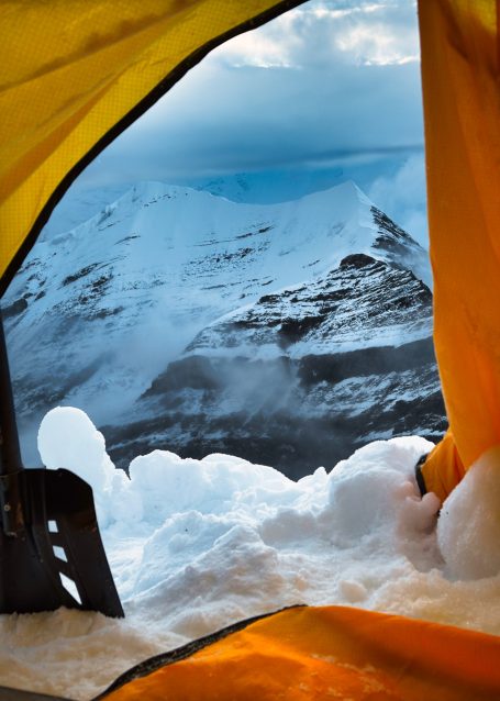 Blick aus einem gelben Zelt auf schneebedeckte Berge und eine weite Aussicht.