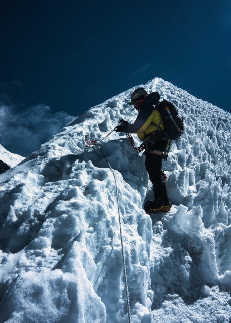 Bergsteiger in gelber Jacke erklimmt schneebedeckten Gipfel unter blauem Himmel.