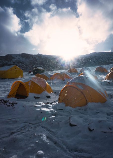 Gelbe Zeltgruppe im Schnee, mit Sonnenstrahlen hinter der bergigen Landschaft.