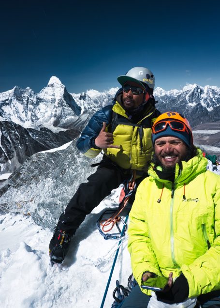 Zwei Bergsteiger posieren auf einem Gipfel mit schneebedeckten Bergen im Hintergrund.