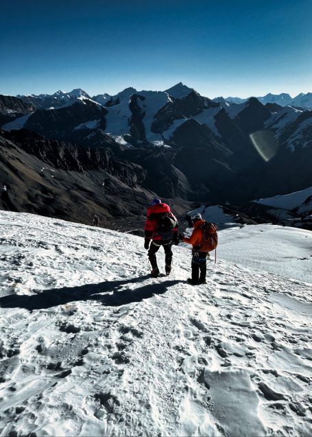 Zwei Personen wandern im Schnee auf einem Berg mit schneebedeckten Gipfeln im Hintergrund.