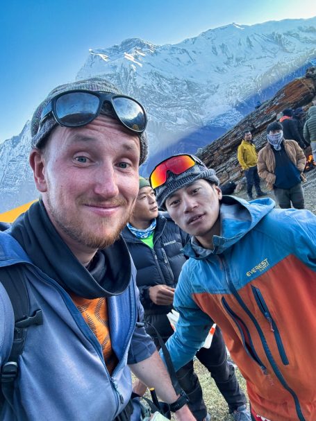 Zwei Männer lächeln in den Bergen, mit schneebedeckten Gipfeln im Hintergrund.