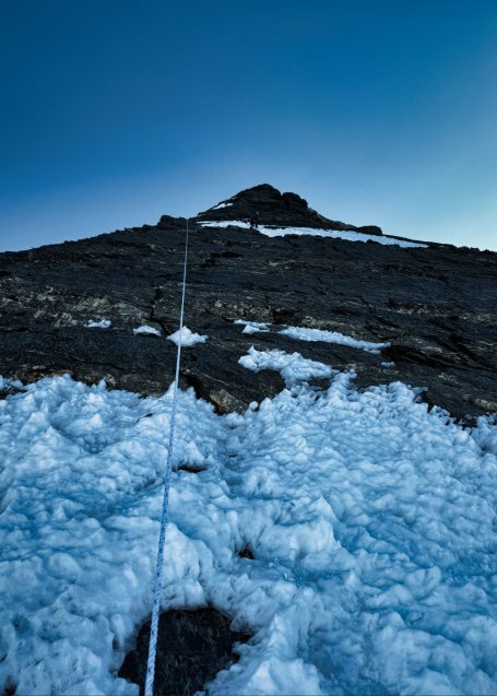 Steiler Berghang mit Schnee und Felsen unter klarem blauen Himmel.