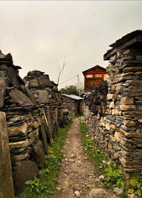 Enge, steinerne Gasse zwischen alten Mauern mit einem Holzhaus im Hintergrund.