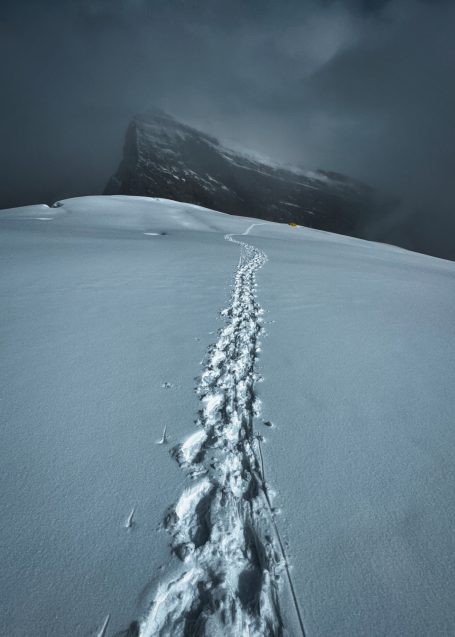 Schneebedeckter Weg mit Fußspuren und einer dramatischen Bergsilhouette im Hintergrund.