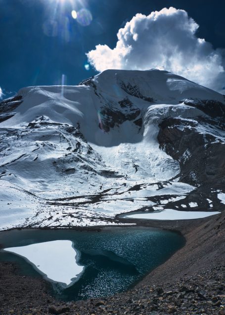 Hoher Berg mit schneebedecktem Gipfel und glitzerndem See im Vordergrund.