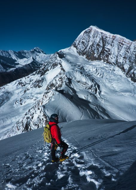 Bergsteiger in roter Jacke steht auf verschneitem Gipfel mit beeindruckendem Ausblick.