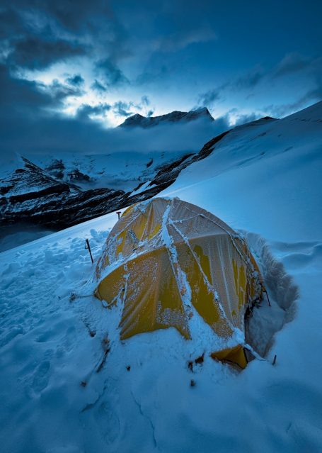 Gelbes Zelt im Schnee unter einem bewölkten Himmel in gebirgiger Umgebung.
