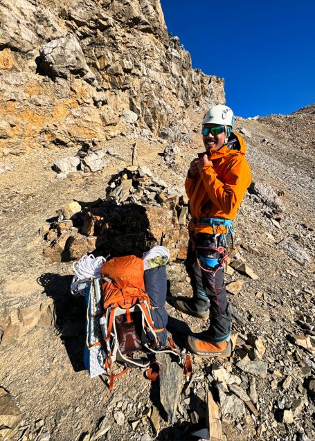 Kletterer in orange Jacke steht an felsiger Bergwand mit Kletterausrüstung.