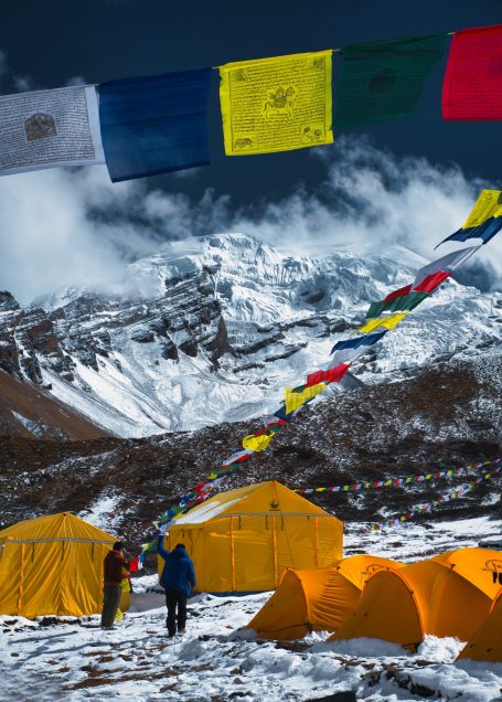 Berglandschaft mit Zelten und bunten Gebetsfahnen vor schneebedeckten Gipfeln.