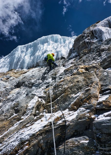 Ein Kletterer erklimmt eine steile Felswand mit Eis oben, unter blauen Himmel.
