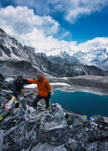Zwei Personen posieren am Berghang mit einem See und schneebedeckten Bergen im Hintergrund.