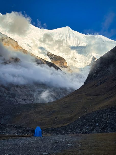 Blauer Zelt in einer bergigen Landschaft mit schneebedeckten Gipfeln und Wolken.