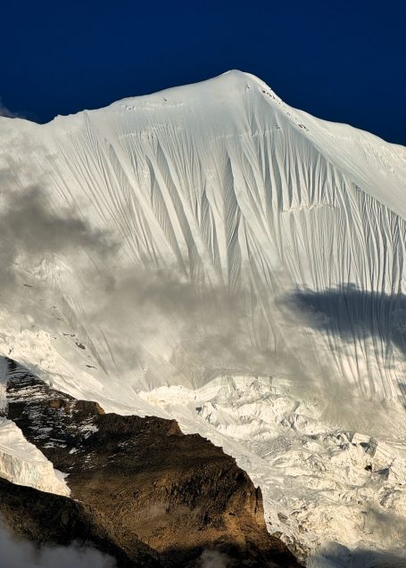 Eine schneebedeckte Bergspitze mit Wolken im Vordergrund und klarem Himmel im Hintergrund.