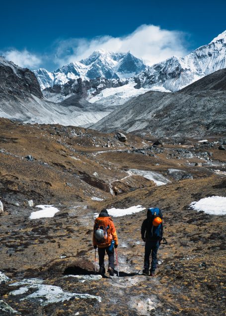 Zwei Wanderer mit Rucksäcken in einer gebirgigen, schneebedeckten Landschaft.