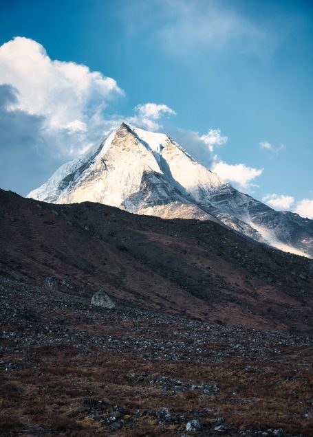 Hoher, schneebedeckter Berg mit Wolken und felsigem Gelände im Vordergrund.
