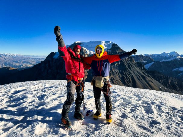 Zwei Bergsteiger jubeln auf dem Gipfel, umgeben von schneebedeckten Bergen und blauem Himmel.