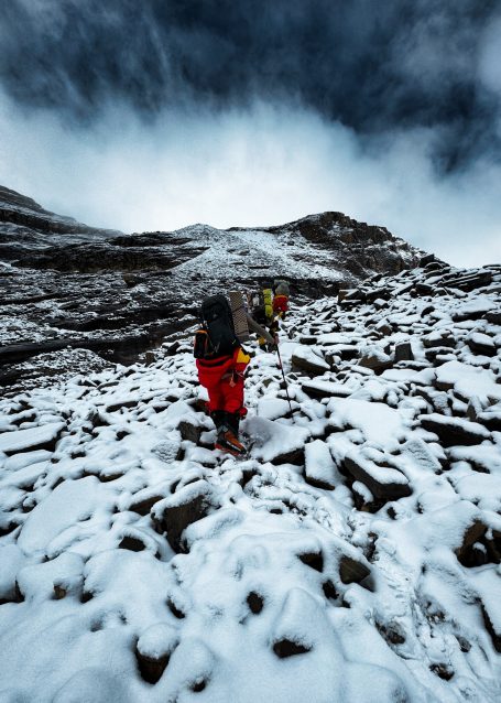 Zwei Personen wandern über einen schneebedeckten, steinigen Berghang unter bewölktem Himmel.