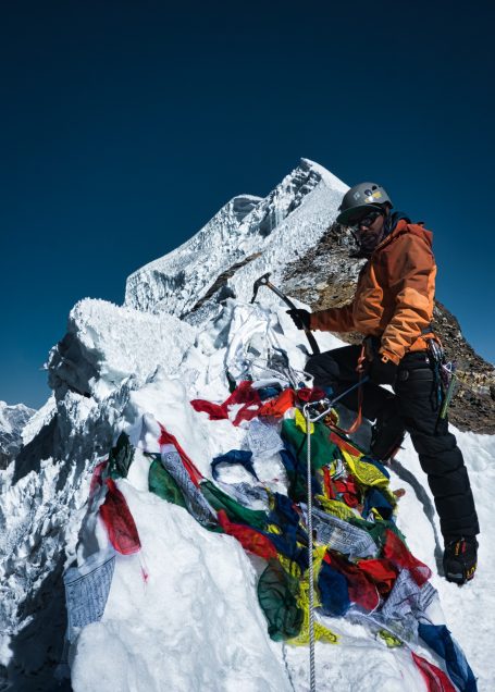 Bergsteiger in orangeem Anzug auf einem schneebedeckten Gipfel mit bunten Bannern.
