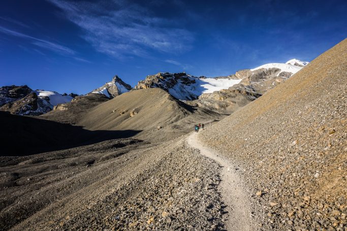 Schmaler Pfad zwischen steinigen Hügeln mit schneebedeckten Bergen im Hintergrund.