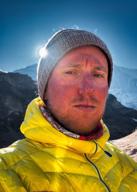 Mann mit gelber Jacke und grauer Mütze vor einem bergigen Hintergrund.