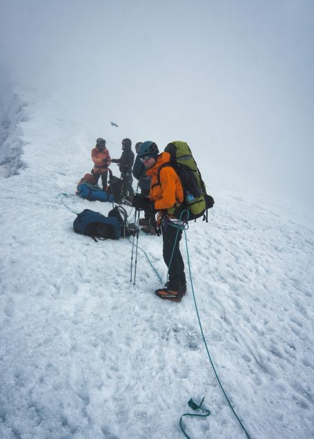 Bergsteiger in orange Jacke auf schneebedecktem Gelände in nebliger Umgebung.