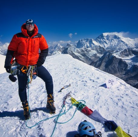 Bergsteiger in orangefarbener Jacke auf einem schneebedeckten Gipfel mit Aussicht auf Berge.