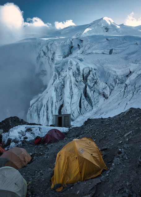 Zelte auf felsigem Gelände mit schneebedeckten Bergen und Wolken im Hintergrund.