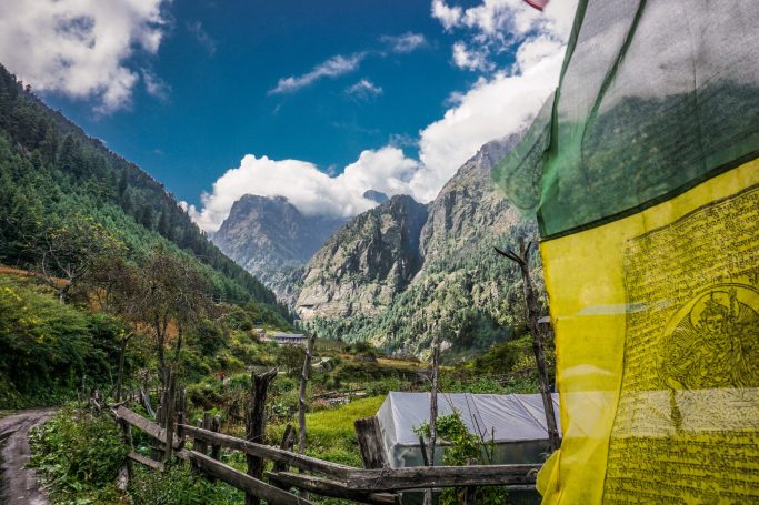Berglandschaft mit bewaldeten Hügeln, klarer Himmel und bunten Gebetsfahnen im Vordergrund.