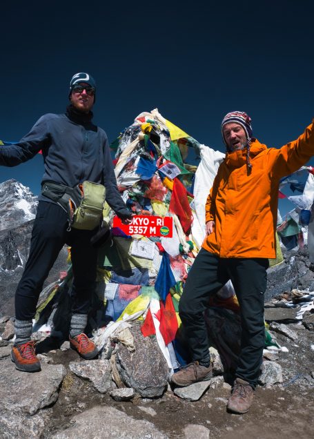 Zwei Bergsteiger posieren vor einem bunten Gipfel mit Gebetsfahnen.