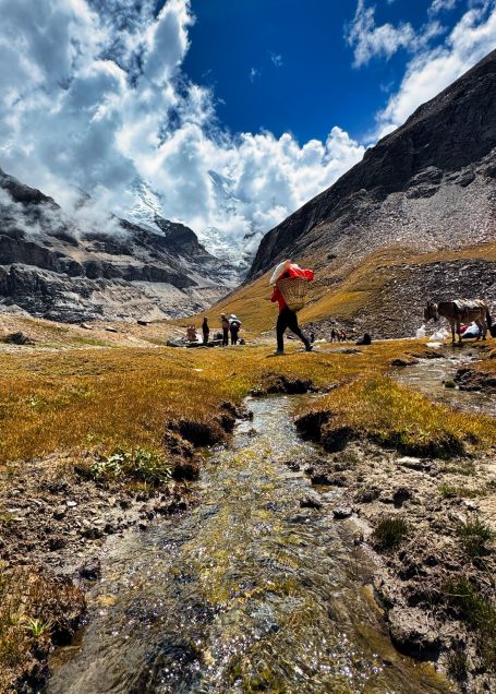 Wanderer mit rotem Rucksack am Rande eines Bachs in den Bergen unter bewölktem Himmel.