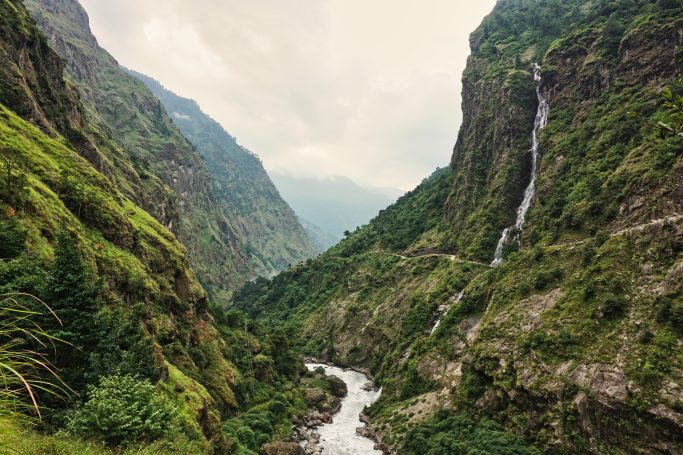 Schmale Schlucht mit Fluss und Wasserfall, umgeben von grünen Bergen und bewölktem Himmel.